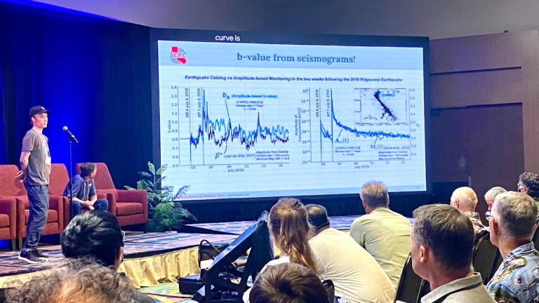 A person on a stage presenting their poster at a SCEC annual meeting in front of a large screen